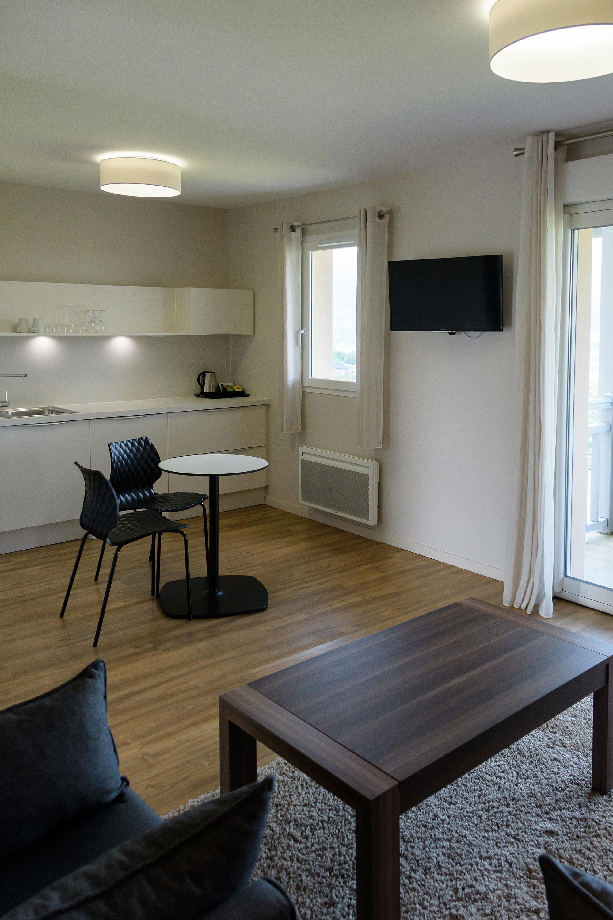 A modern living room with a sofa, a coffee table, a dining table with chairs, a kitchen area, and a TV mounted on the wall.