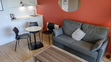 Un espace de vie compact comprenant un coin cuisine, un canapé gris agrémenté de coussins, un miroir rond et une petite table ronde.
