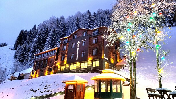 A large, multi-story building with a prominent entrance and a small adjacent structure, surrounded by snow-covered trees and a clear sky. - Ayderoom Otel (Camlihemsin)
