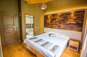 Una habitación de hotel con piso de madera, una cama con cubrecama a rayas, cabecera de madera, mesita de noche e lámpara colgante.