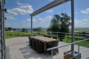 A terrace with a wooden table and wicker chairs, overlooking a green landscape with trees and houses.