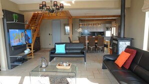 Un salon moderne avec un canapé en cuir noir, une table basse en verre et un téléviseur posé sur un meuble. Un escalier mène à l’étage supérieur.