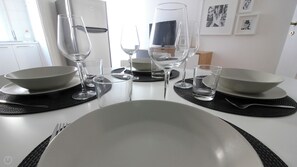 A dining table set with plates, bowls, and wine glasses, with a refrigerator and framed pictures in the background.