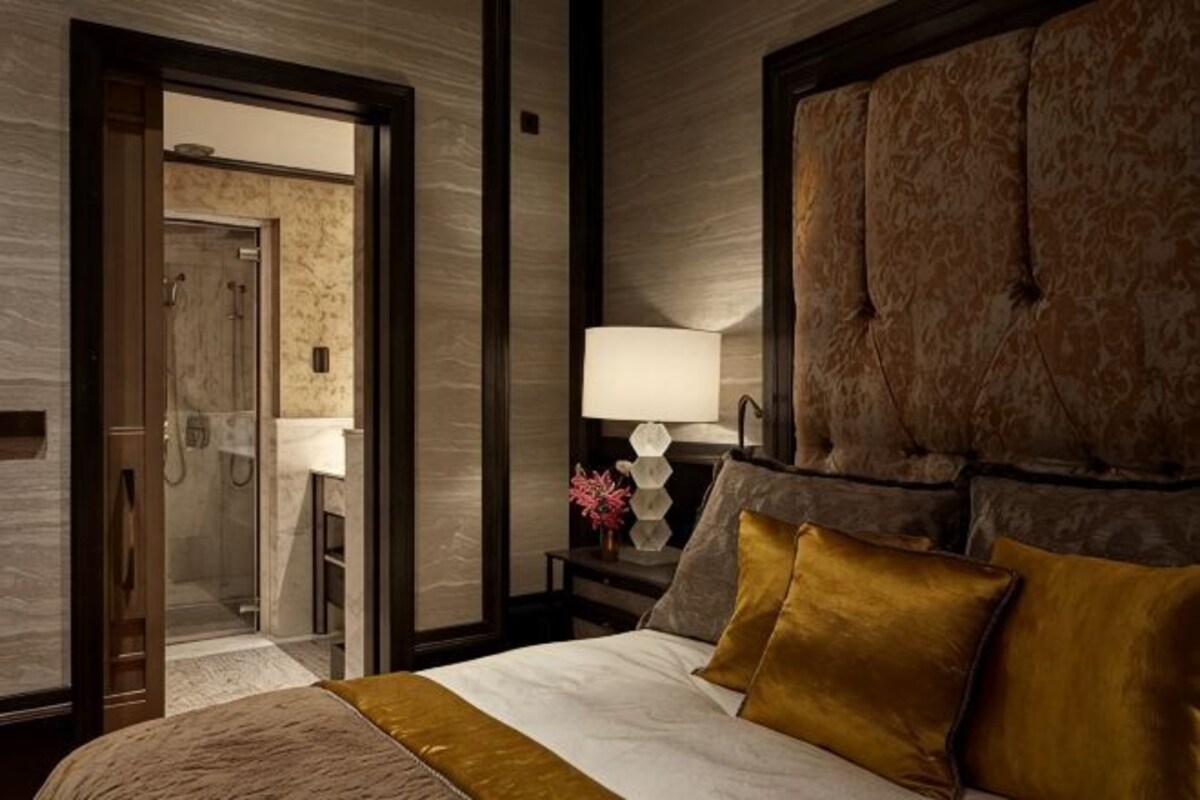 Habitación de hotel con una cama grande, un cabecero, una mesita de noche con una lámpara y un baño visible a través de una puerta abierta.