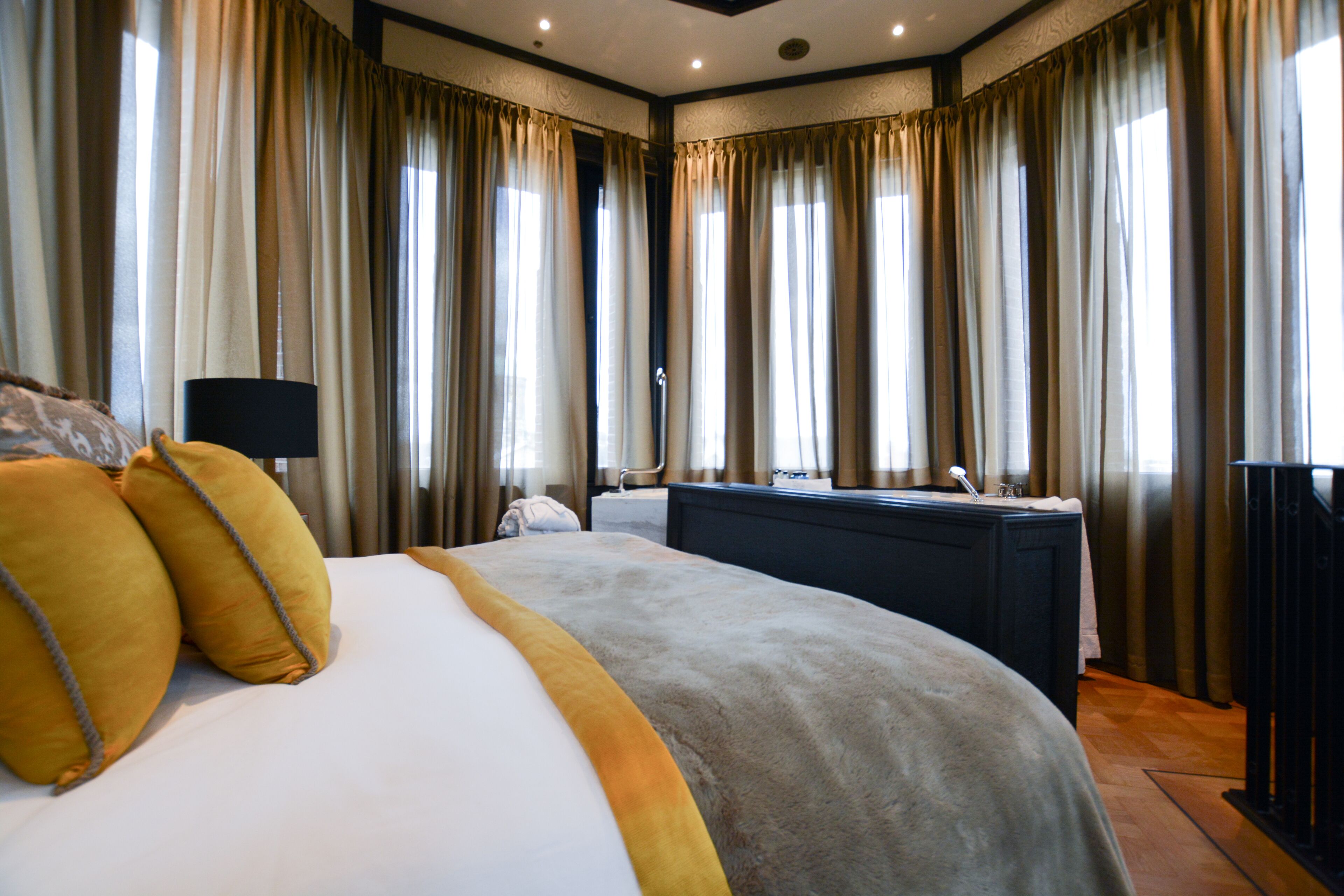 A hotel room with a large bed, a desk with a lamp, and a view of the city through the windows.