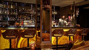 Zona de bar con estanterías llenas de botellas de licor, un barman preparando una bebida y asientos con un sofá y sillones cómodos.