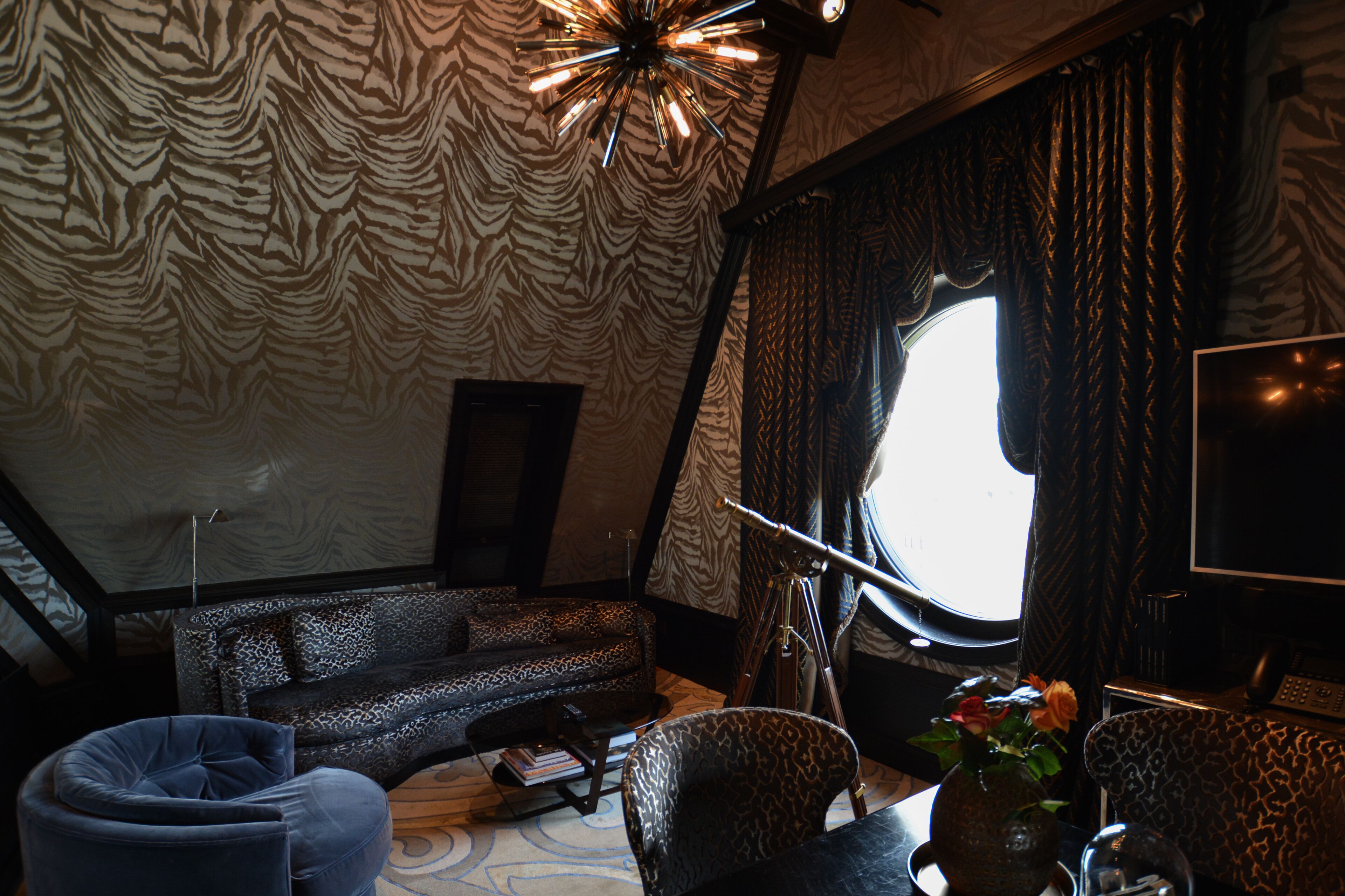 Una habitación con techo estampado de cebra, una lámpara moderna, un sofá, una silla, una mesa con un jarrón de flores y un ventanal con cortinas.