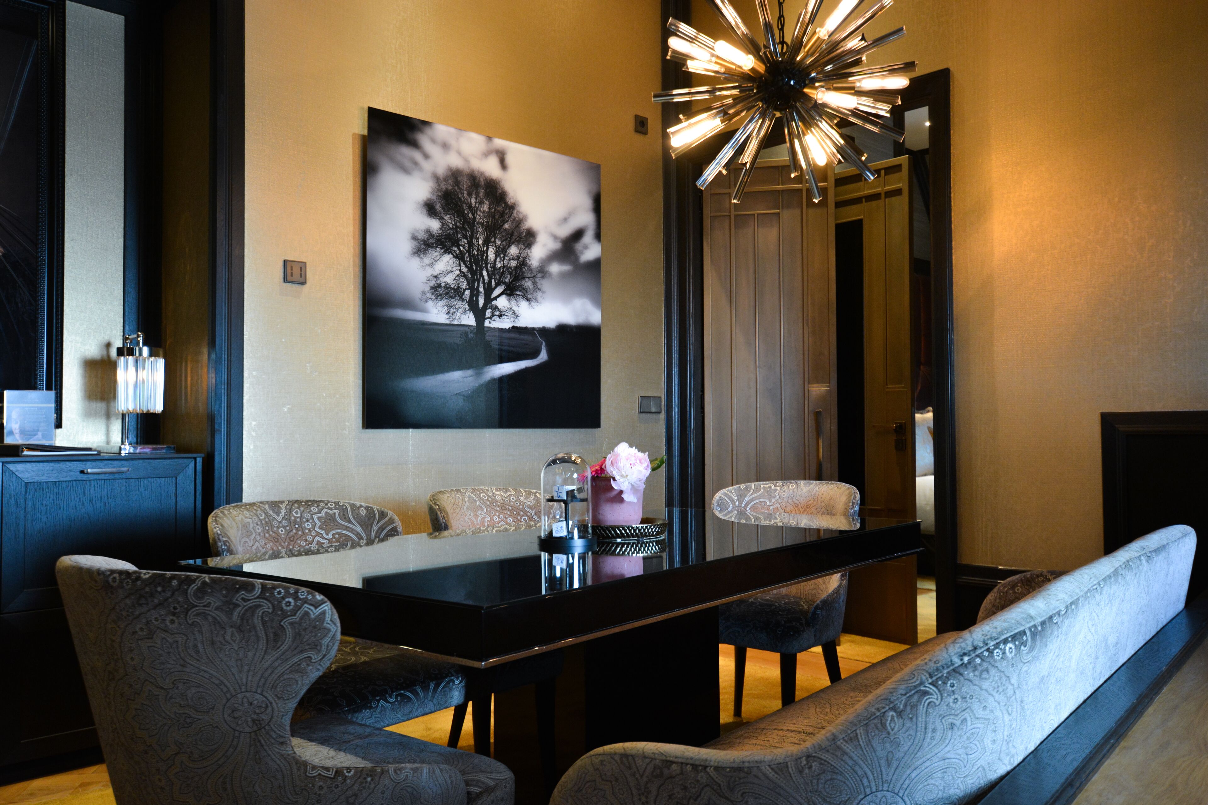 A modern dining area with a long table, patterned chairs, and a wall-mounted artwork of a tree.