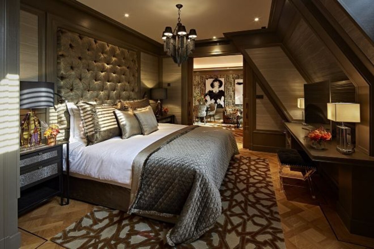 A bedroom with a large bed, a chandelier, a desk with a lamp, and a patterned rug.