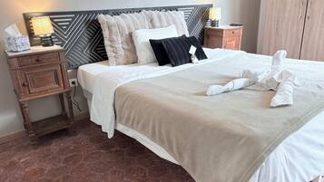 A neatly made bed with a beige blanket, white pillows, and a black pillow. There is a wooden nightstand with a lamp and a tissue box. The room has a wooden headboard and a wooden wardrobe.