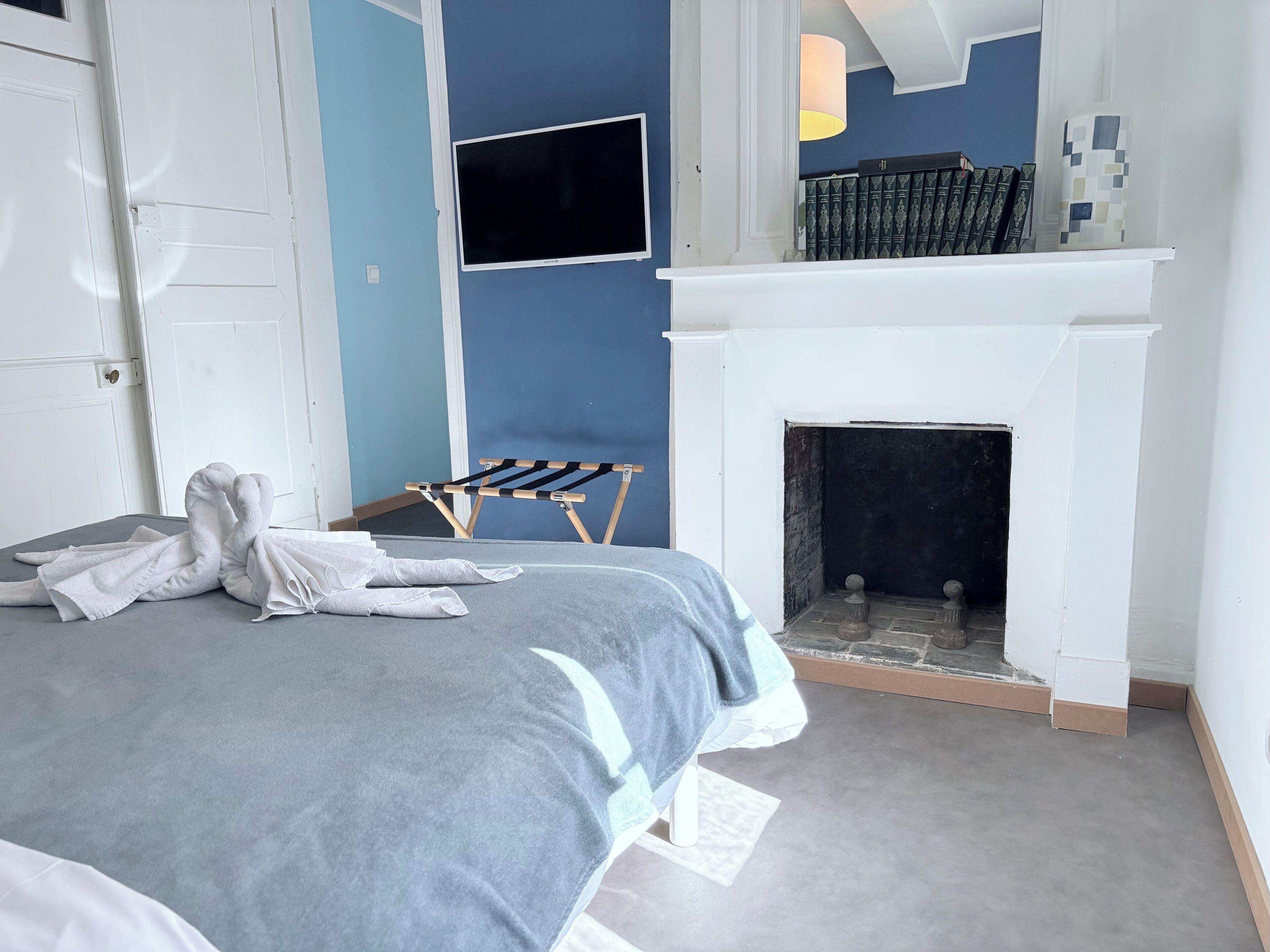 A neatly made bed with white towels, a small wooden stool, a fireplace, and a bookshelf with books.