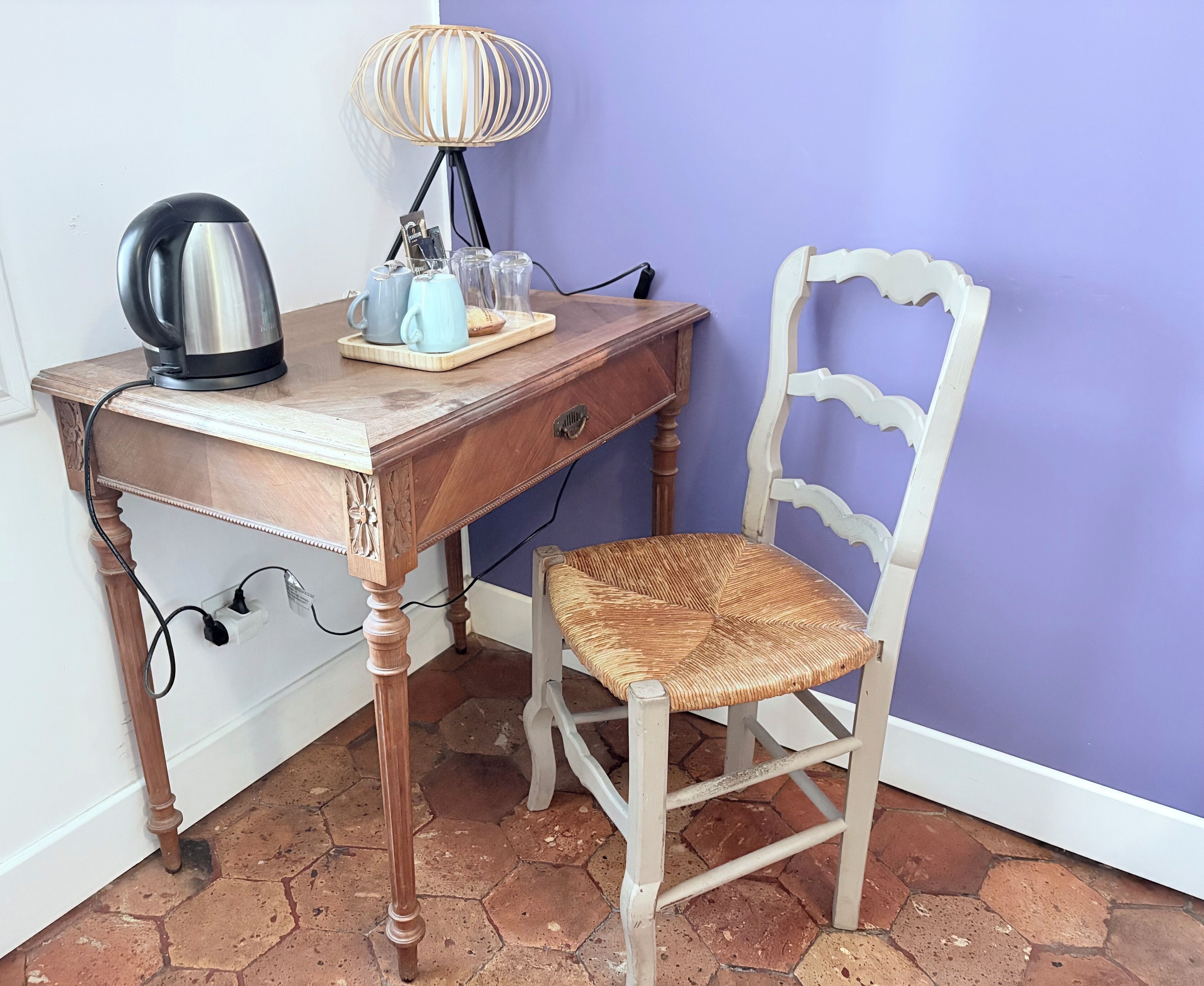 Un bureau en bois, avec une bouilloire, une lampe et une chaise dont l’assise est en osier tressé.