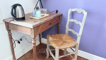Un bureau en bois, avec une bouilloire, une lampe et une chaise dont l’assise est en osier tressé.