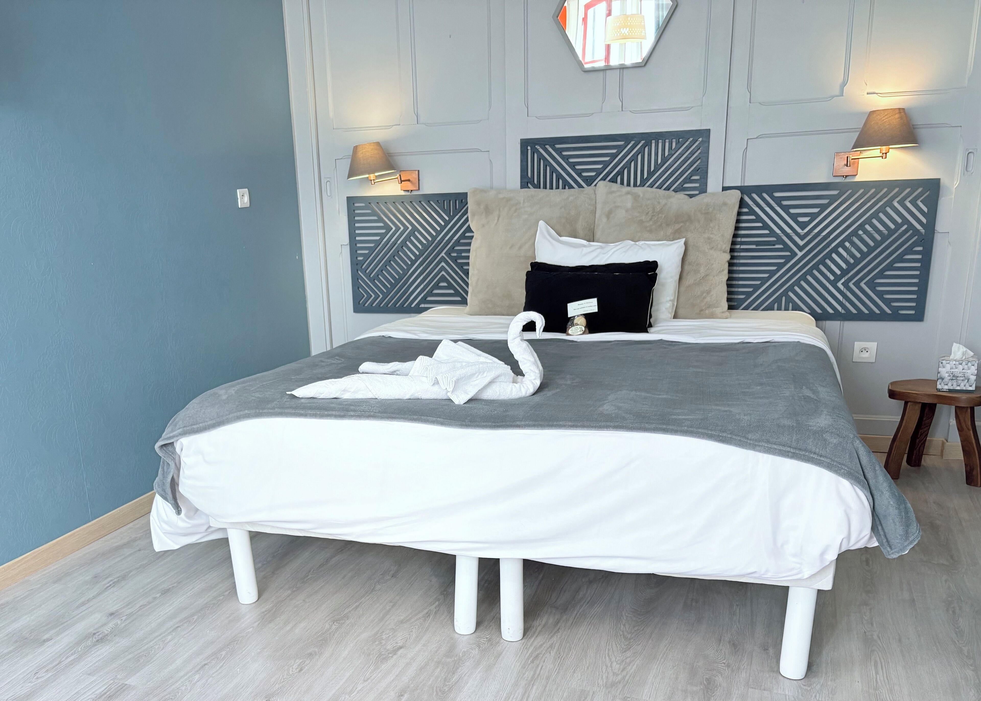 A neatly made bed with a grey and white duvet, a decorative swan towel, and a small bedside table with a tissue box.