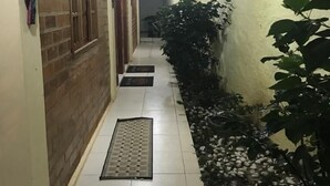 Un pasillo estrecho con suelo de baldosas, un felpudo metálico en la entrada y una planta en maceta a la derecha.