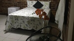 Una habitación con una cama, una mesita con un florero, un ventilador de techo, una pared de ladrillo y un cartel.