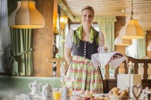 Uma pessoa vestida com traje tradicional, parada em uma mesa com uma refeição montada, incluindo um bule, xícaras e pão.
