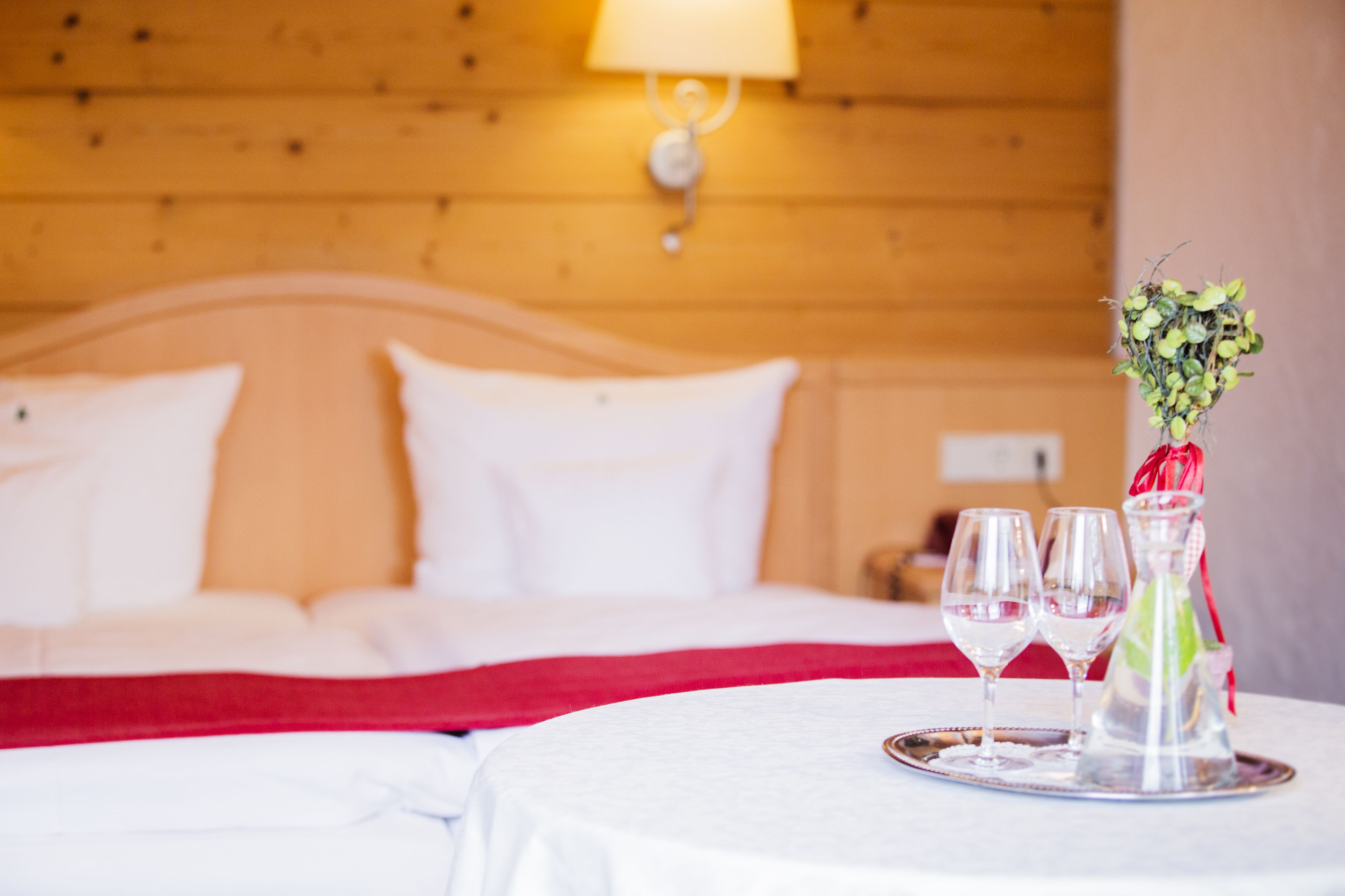 Ein ordentlich bezogenes Bett mit weißen Laken und einer roten Tagesdecke, ein Nachttisch mit zwei Gläsern und einer Pflanze in einer Vase, sowie eine Holzwand mit einer Lampe.