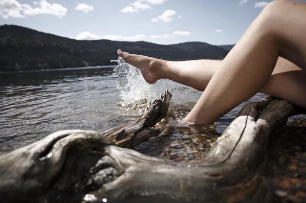 Eine Person sitzt auf einem Baumstamm im Wasser, mit Bergen im Hintergrund.