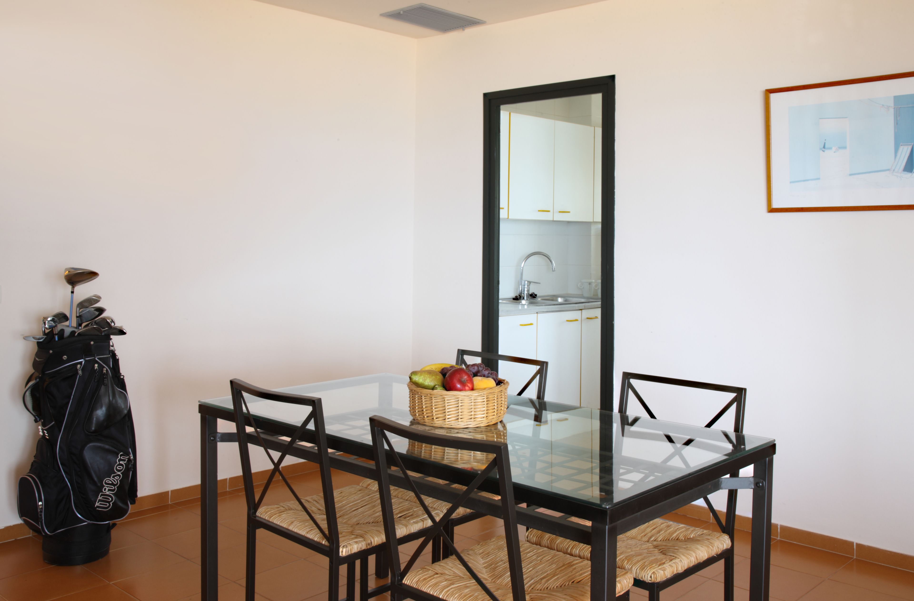 Een eetgedeelte met een glazen tafel, vier stoelen, een rieten mand met fruit en een golfbag.
