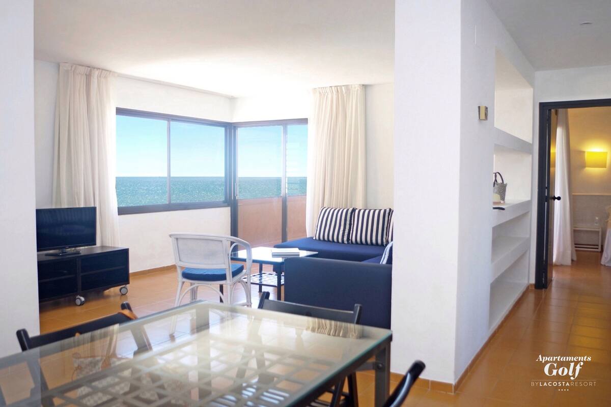 A modern hotel room with a large window, a sofa, a TV, and a glass table.