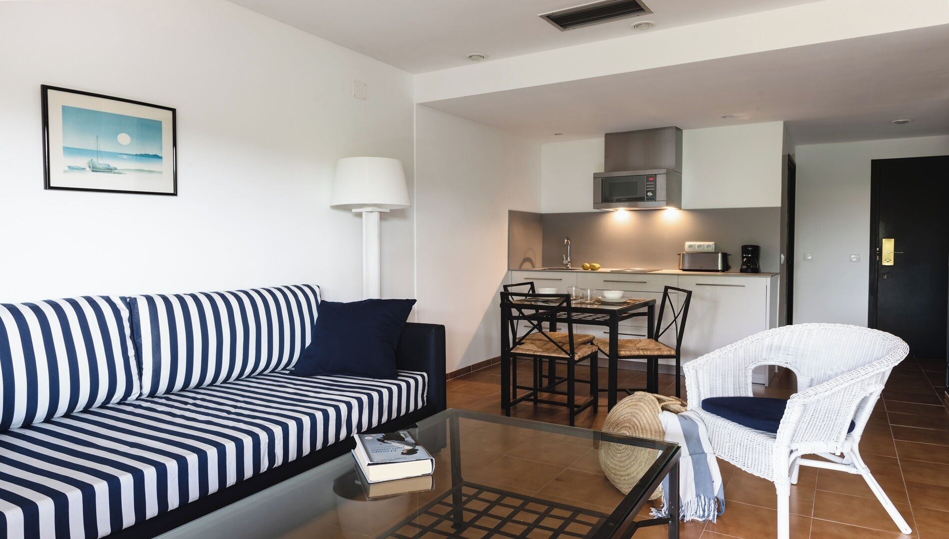 En moderne stue med en stribet sofa, et glasbord, en hvid fletstol og et køkkenområde med en mikrobølgeovn og et spisebord.