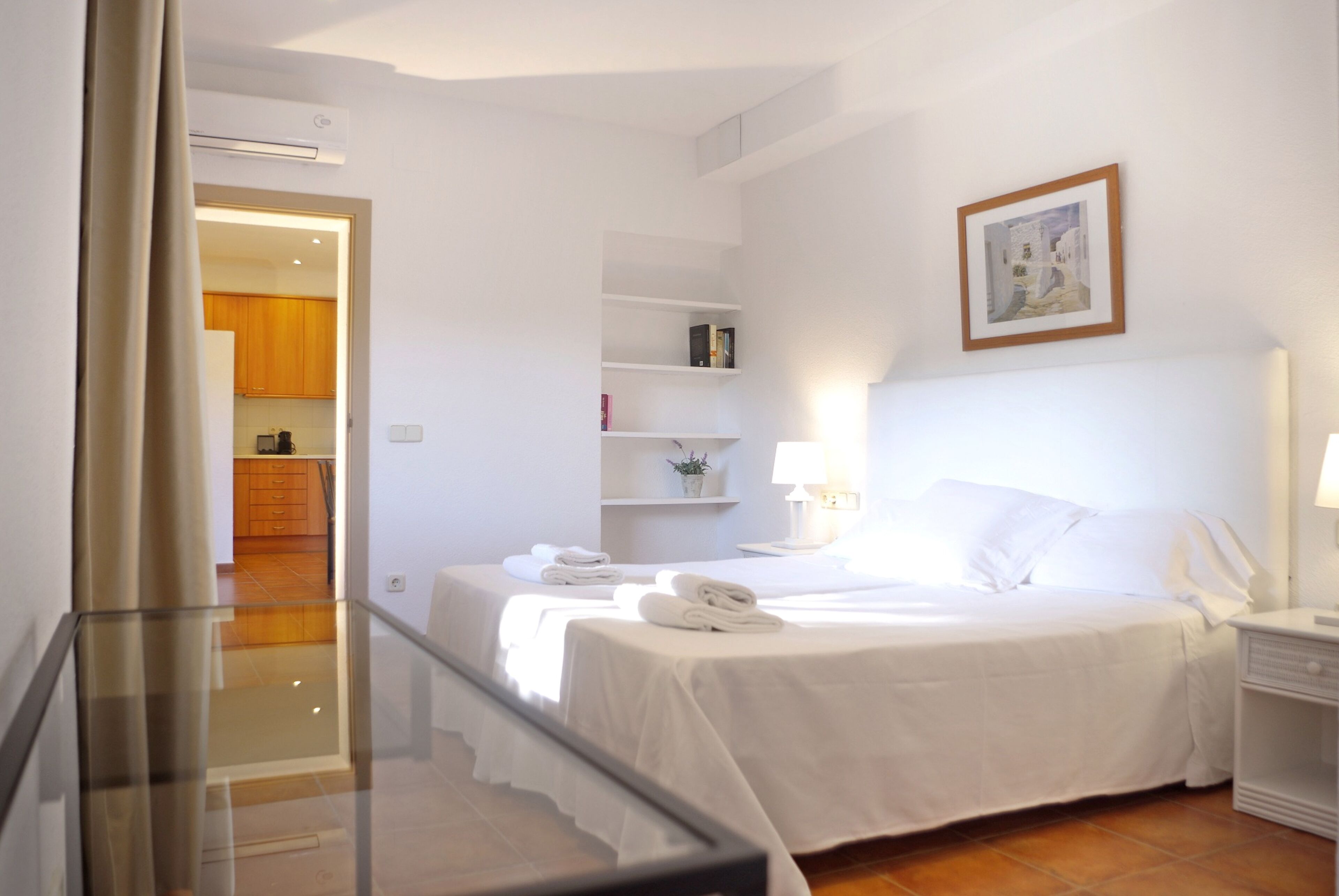 A hotel room with a bed, bedside tables, a lamp, a shelf with books, and a painting on the wall.