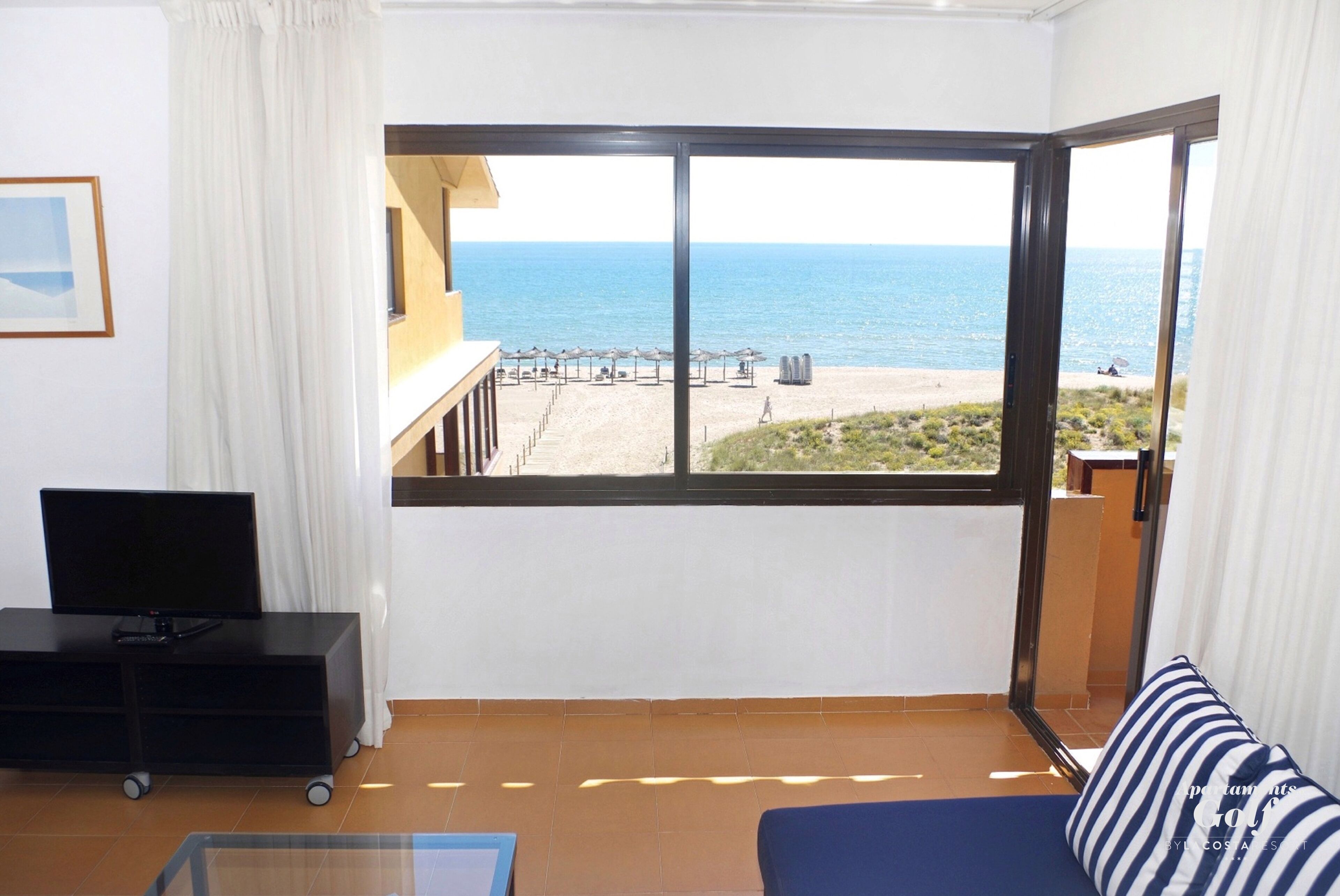A room with a large window offering a view of the beach and ocean. There is a sofa, a TV on a stand, and a glass table.