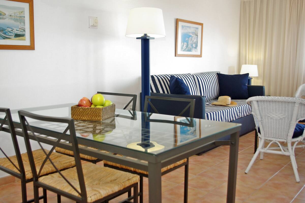 A dining area with a glass table, wicker chairs, a basket of fruit, and a striped sofa with pillows.