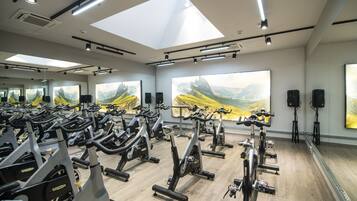 A gym with multiple stationary bikes arranged in rows, large windows with mountain landscape paintings, and ceiling lights.