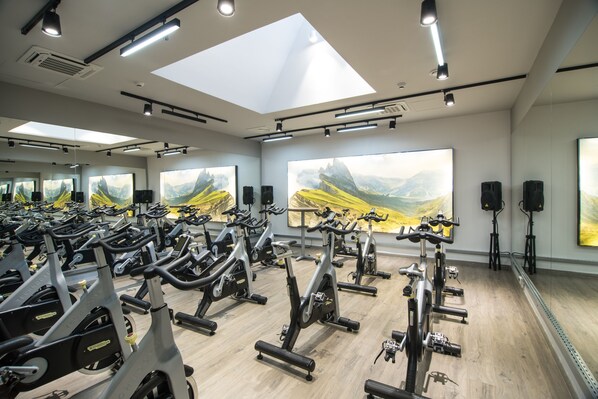 A gym with multiple stationary bikes arranged in rows, large windows with mountain landscape paintings, and ceiling lights.