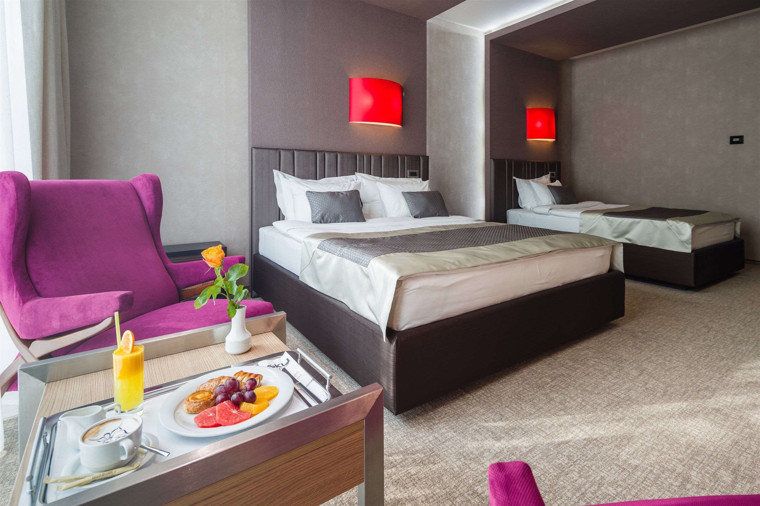 A modern hotel room with a large bed, a smaller bed, a bright red wall lamp, a purple armchair, and a breakfast tray with fruit and coffee.