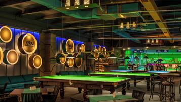 Um salão de bilhar com várias mesas de bilhar, mesas com feltro verde e uma área de bar iluminada em verde.