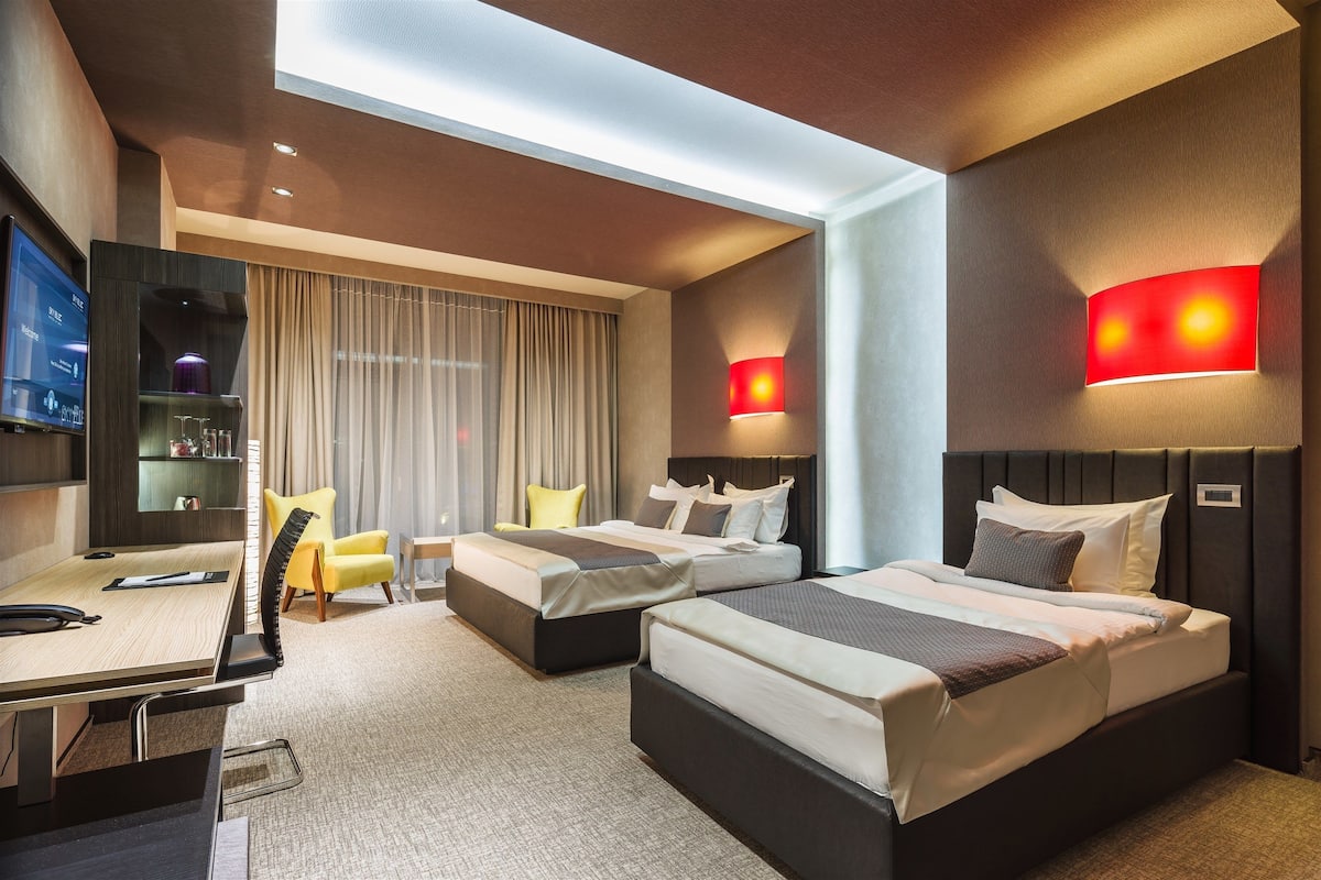Quarto de hotel com duas camas, uma mesa, uma cadeira, uma televisão e uma janela grande com cortinas.