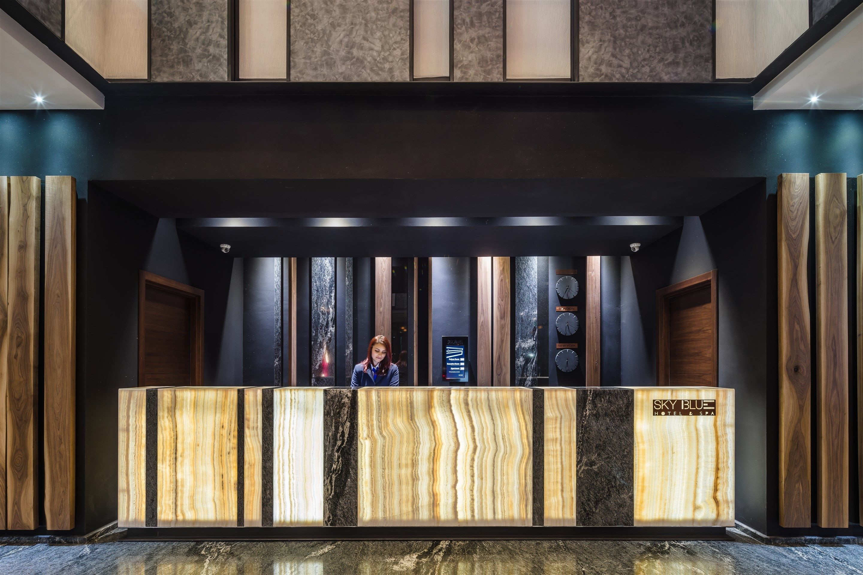 Een receptiebalie met een ontwerp van hout en marmer, een receptionist en een digitaal scherm.