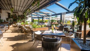 An outdoor dining area with wooden tables and chairs, surrounded by potted plants and a view of a modern building. - Sky Blue Hotel & Spa (Paulestii Noi)