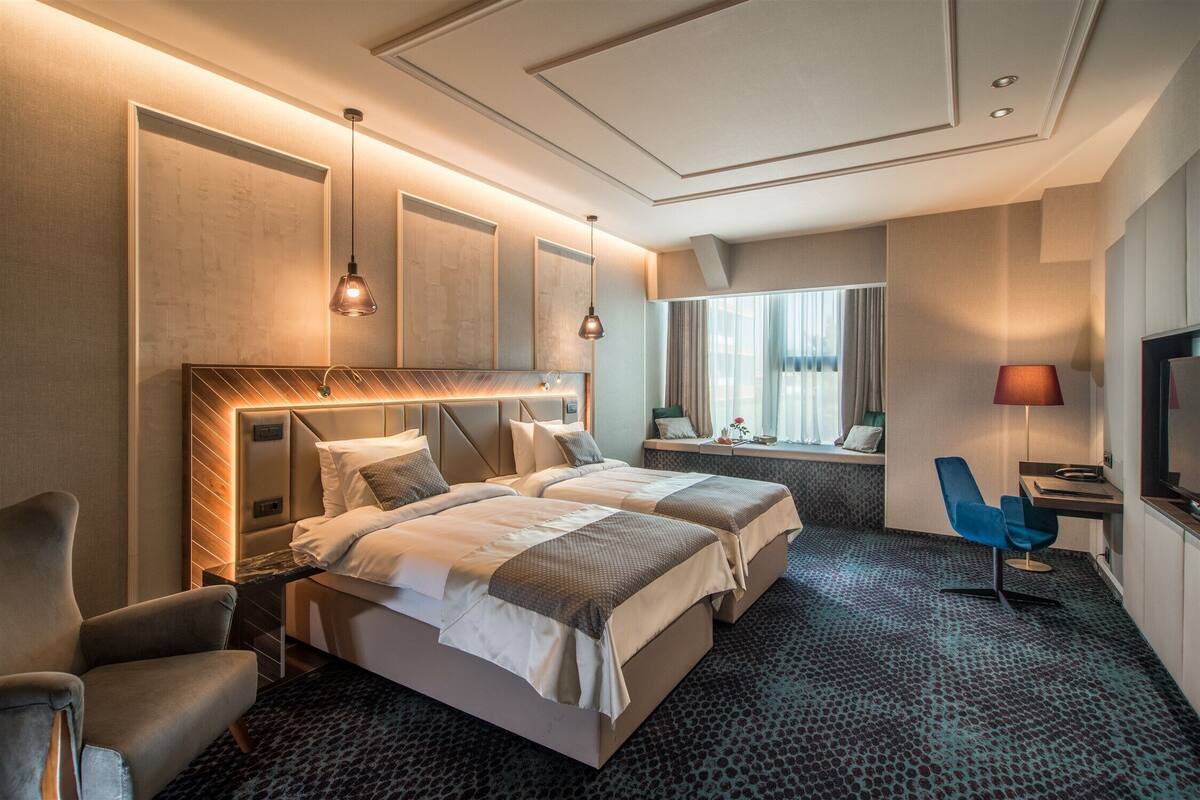 Quarto de hotel com duas camas, uma escrivaninha, uma cadeira, uma TV e uma janela com cortinas.