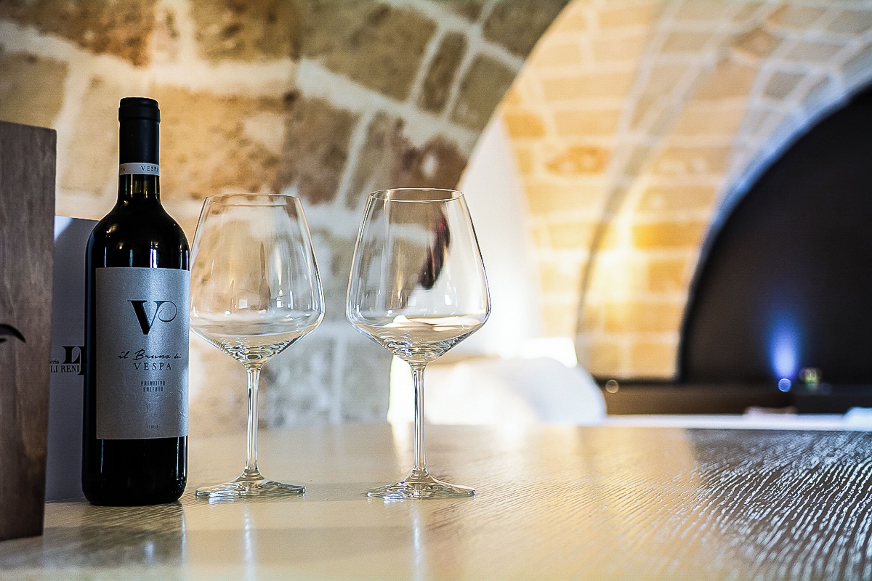 A bottle of wine and two glasses on a table in a stone-walled room.