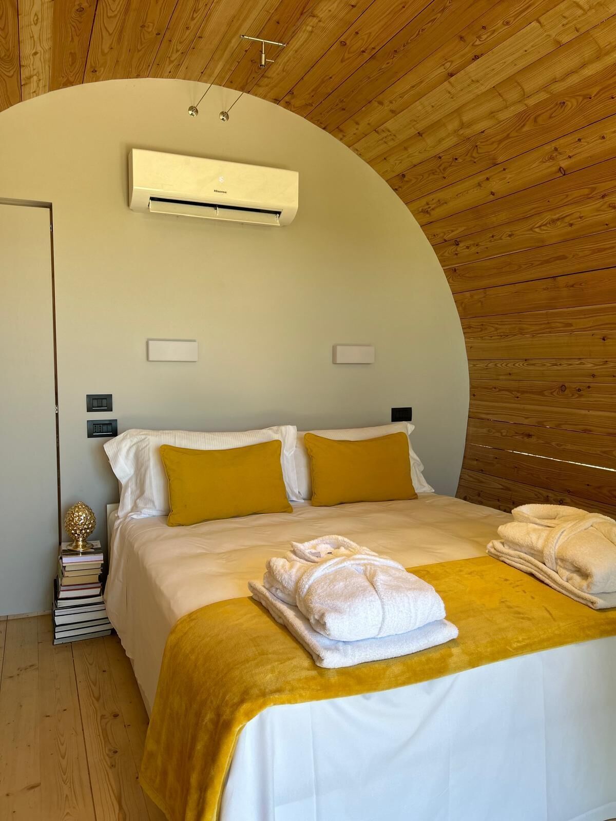 Une chambre à coucher avec un plafond incurvé, un lit avec une literie blanche et jaune moutarde, une table de chevet avec des livres et un appareil de climatisation.