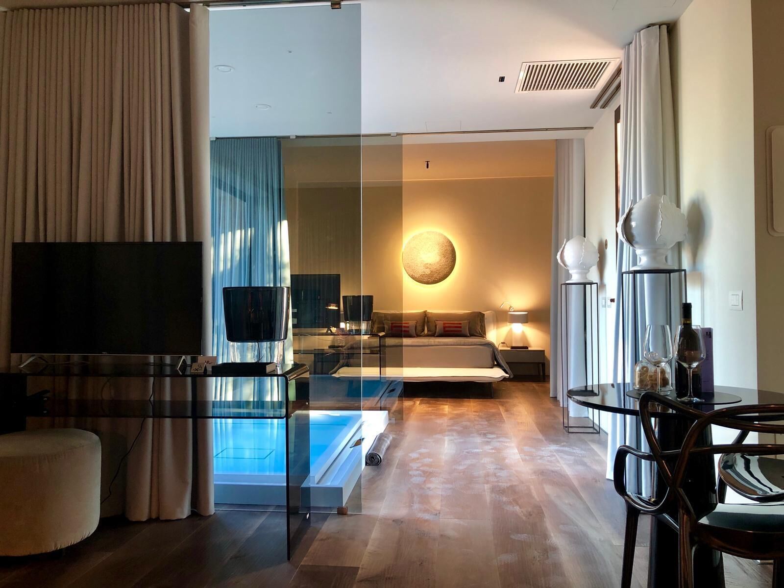 Une chambre d’hôtel moderne dotée d’une cloison en verre, d’un téléviseur à écran plat, d’un canapé, d’une applique murale ronde et d’un coin repas avec une table et des chaises en verre.