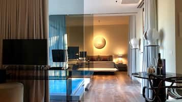 Une chambre d’hôtel moderne dotée d’une cloison en verre, d’un téléviseur à écran plat, d’un canapé, d’une applique murale ronde et d’un coin repas avec une table et des chaises en verre.