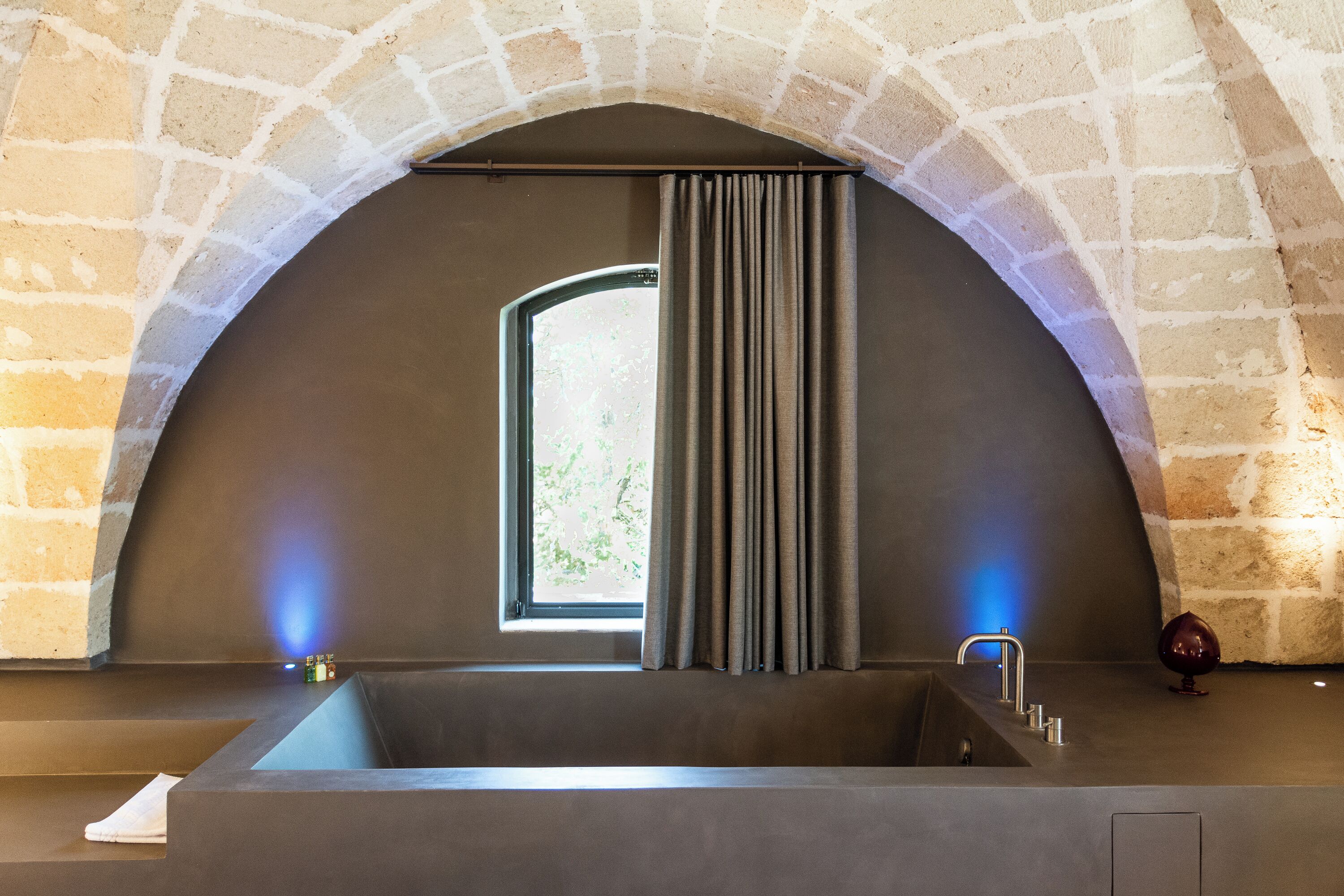 A modern bathroom with a large bathtub, a window with curtains, and a stone archway.