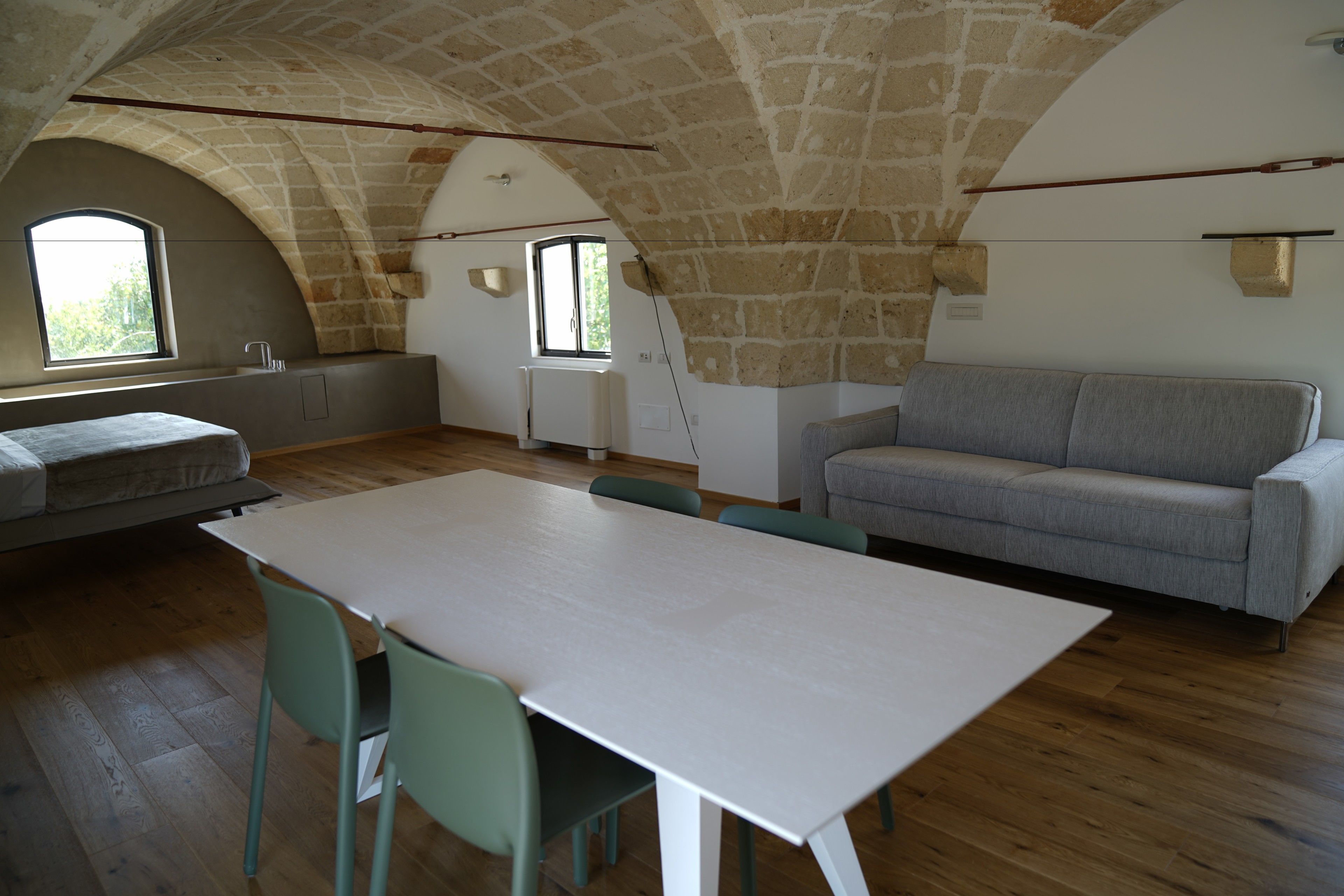 A spacious room with a high arched stone ceiling, a white table with chairs, a grey sofa, and a window providing natural light.