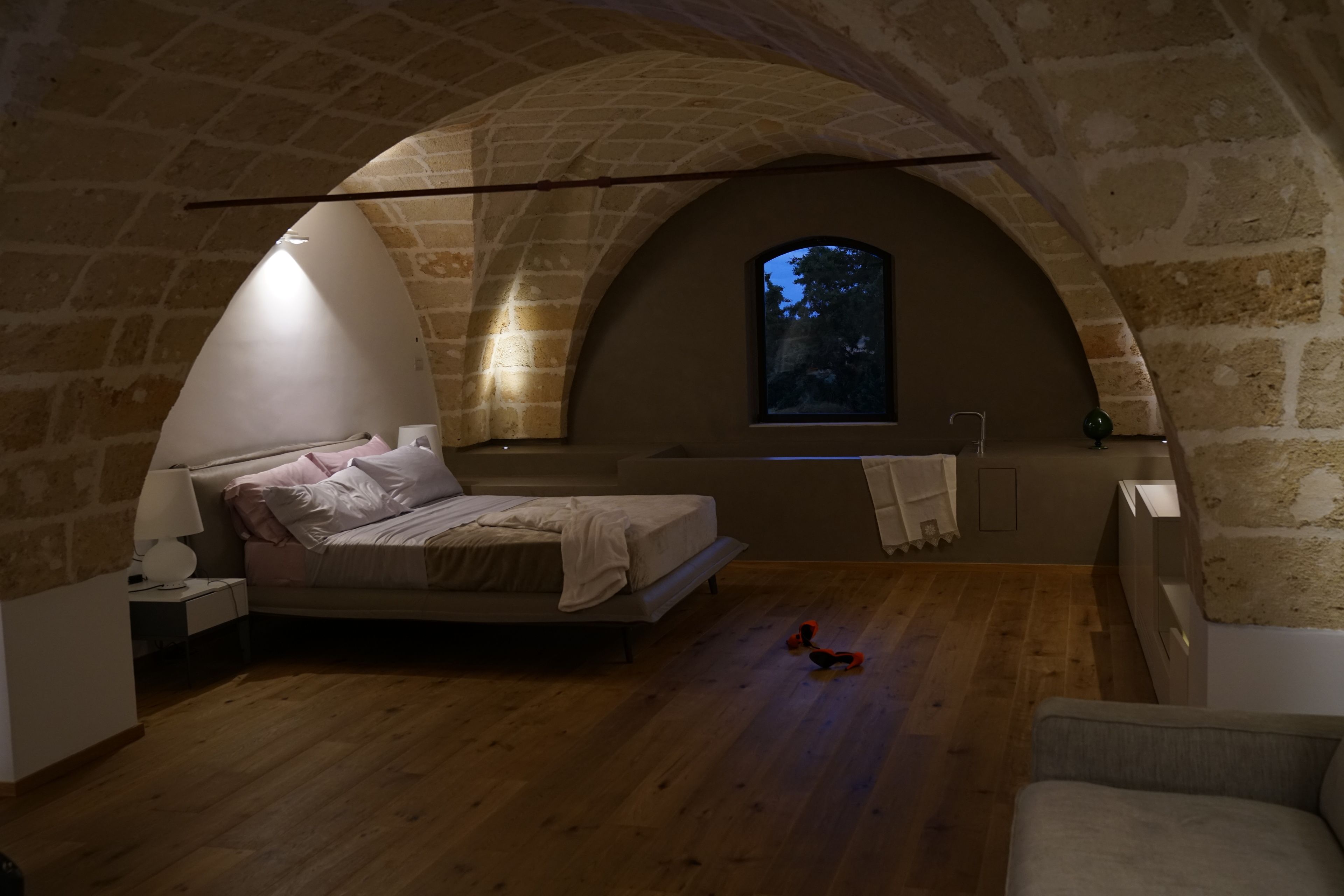 A bedroom with a stone arch ceiling, a bed, a nightstand, a window, and a wooden floor.