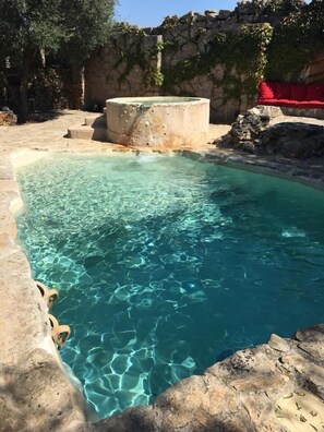 A clear pool with stone steps leading into it, surrounded by a stone wall and vegetation.