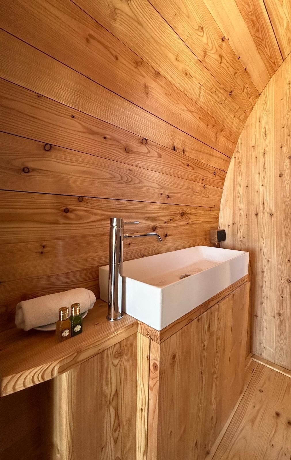 A wooden cabin interior with a built-in white sink, a towel, and two bottles on a shelf.