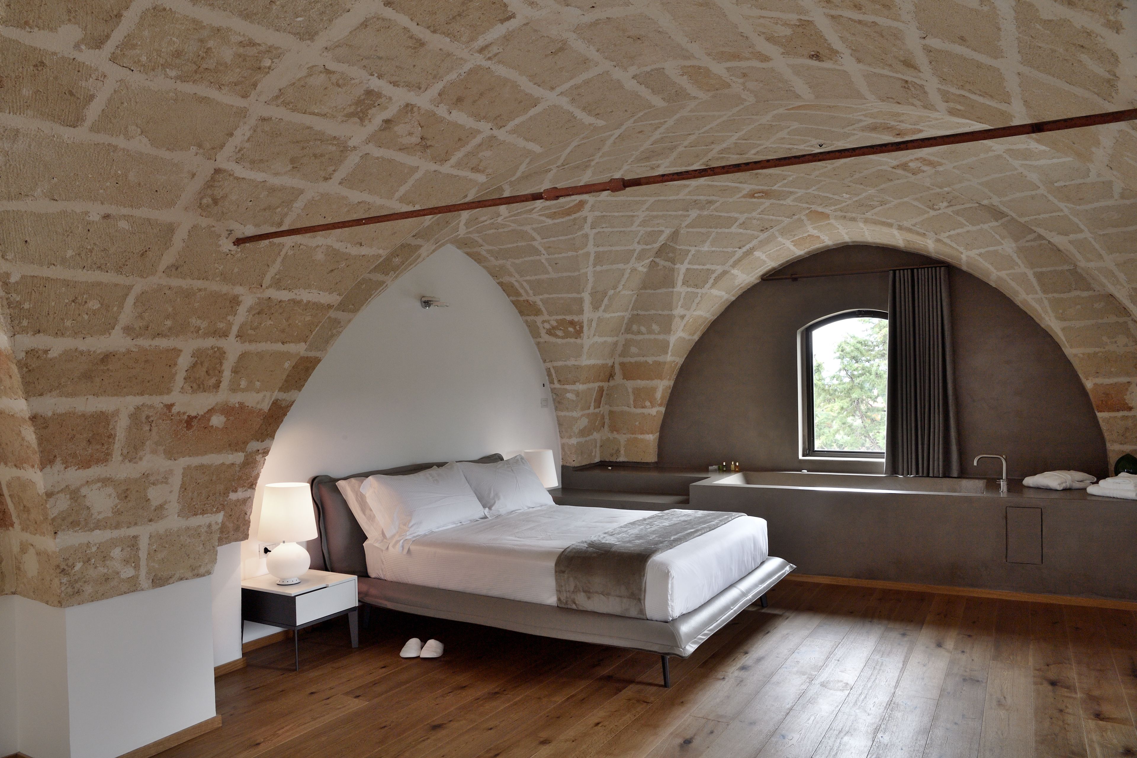 Um quarto com teto abobadado de pedra, uma cama com cabeceira branca, uma mesa de cabeceira com abajur, uma janela com cortinas e um banco embutido com espelho e porta-toalhas.