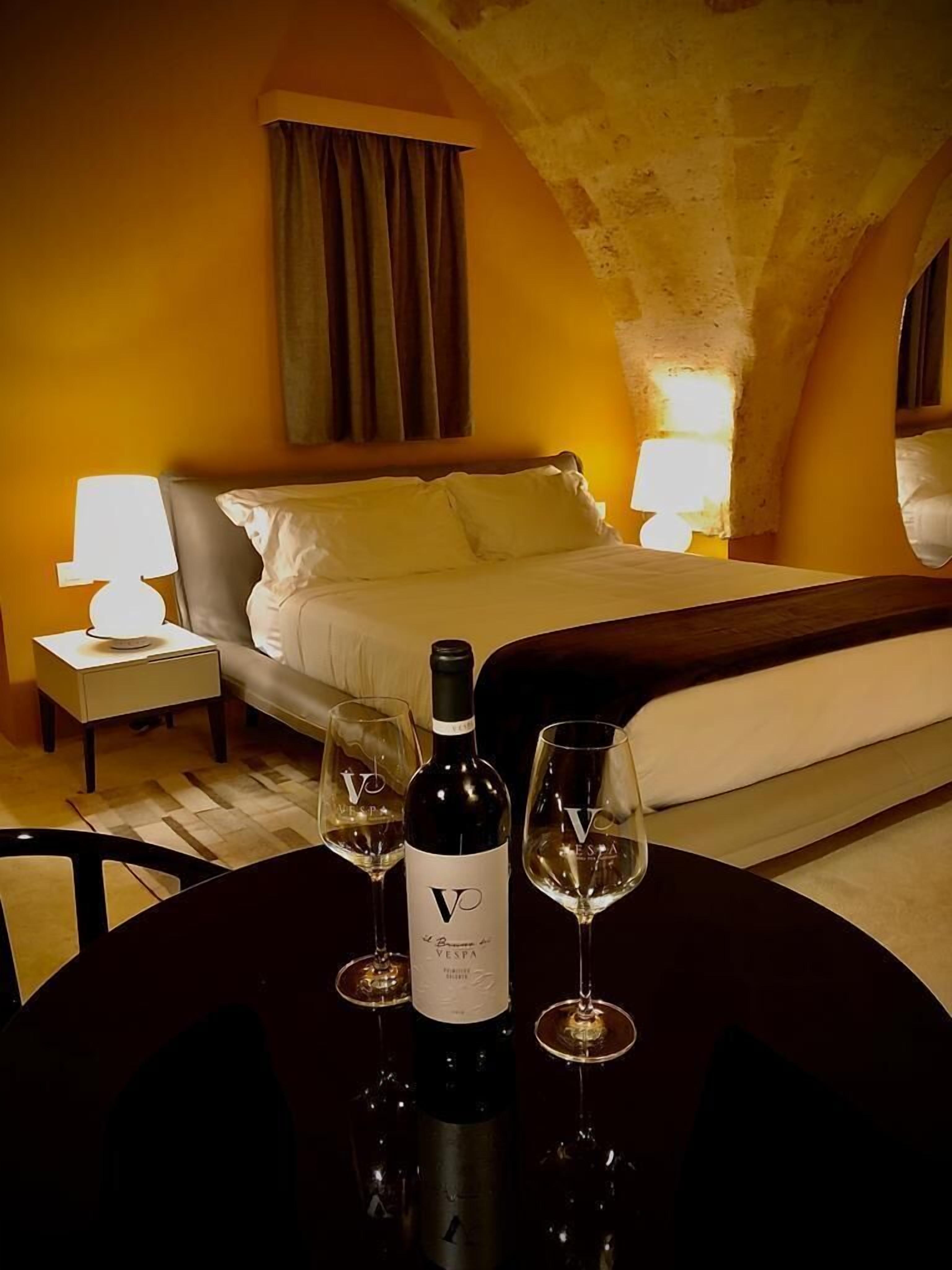 A hotel room with a bed, a table with a bottle of wine and two glasses, and a nightstand with a lamp.