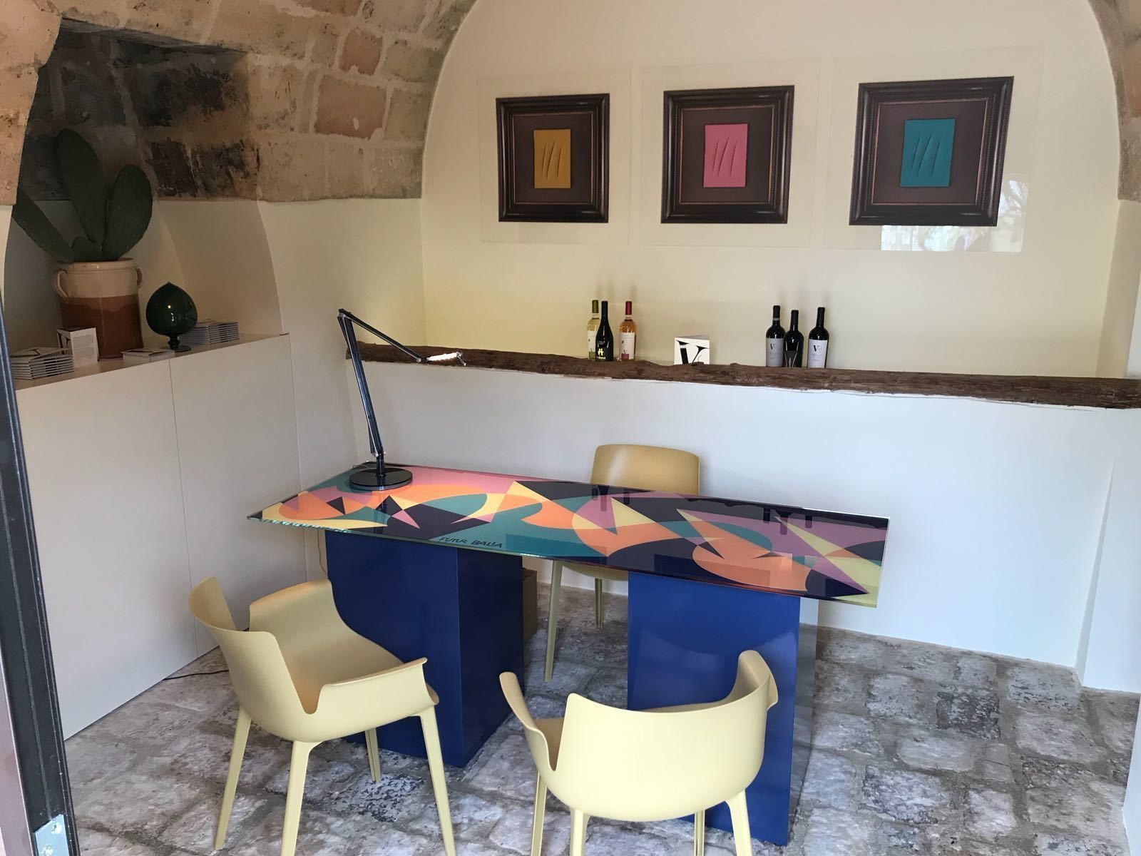 A bar area with a colorful geometric-patterned table, two yellow chairs, a dark desk lamp, and three framed artworks on the wall.