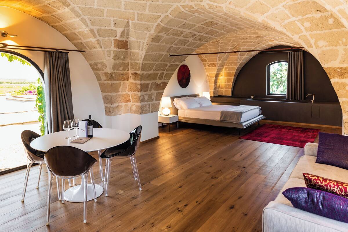 Ein gemütliches Zimmer mit Steindecke, einem runden Esstisch mit Stühlen, einem Bett mit roter Wand und einem Fenster mit Vorhängen.